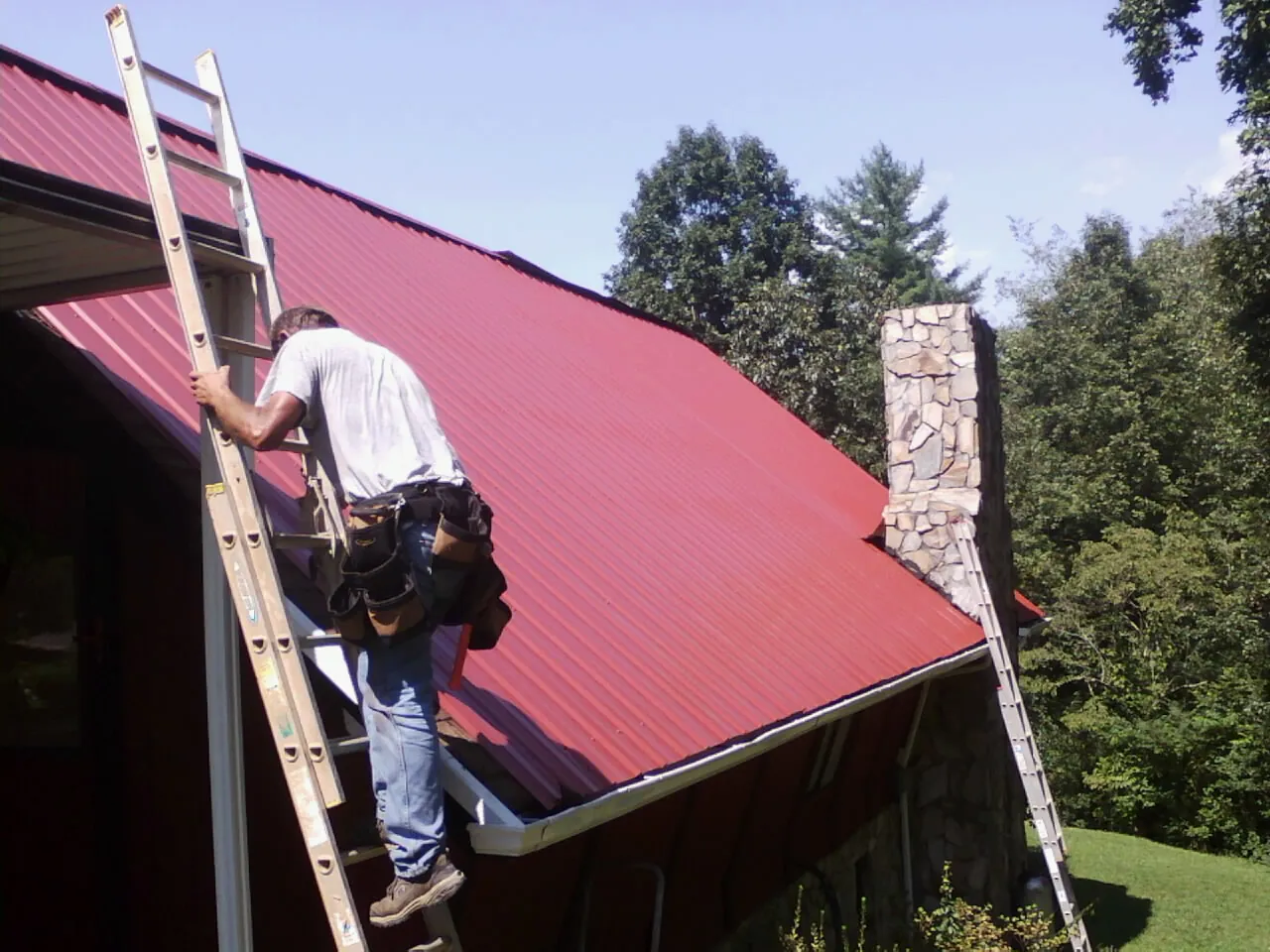 Roofer on a ladder beside a red metal roof and stone chimney — Soffit & Fascia Repair by Roofing Brothers in Toms River